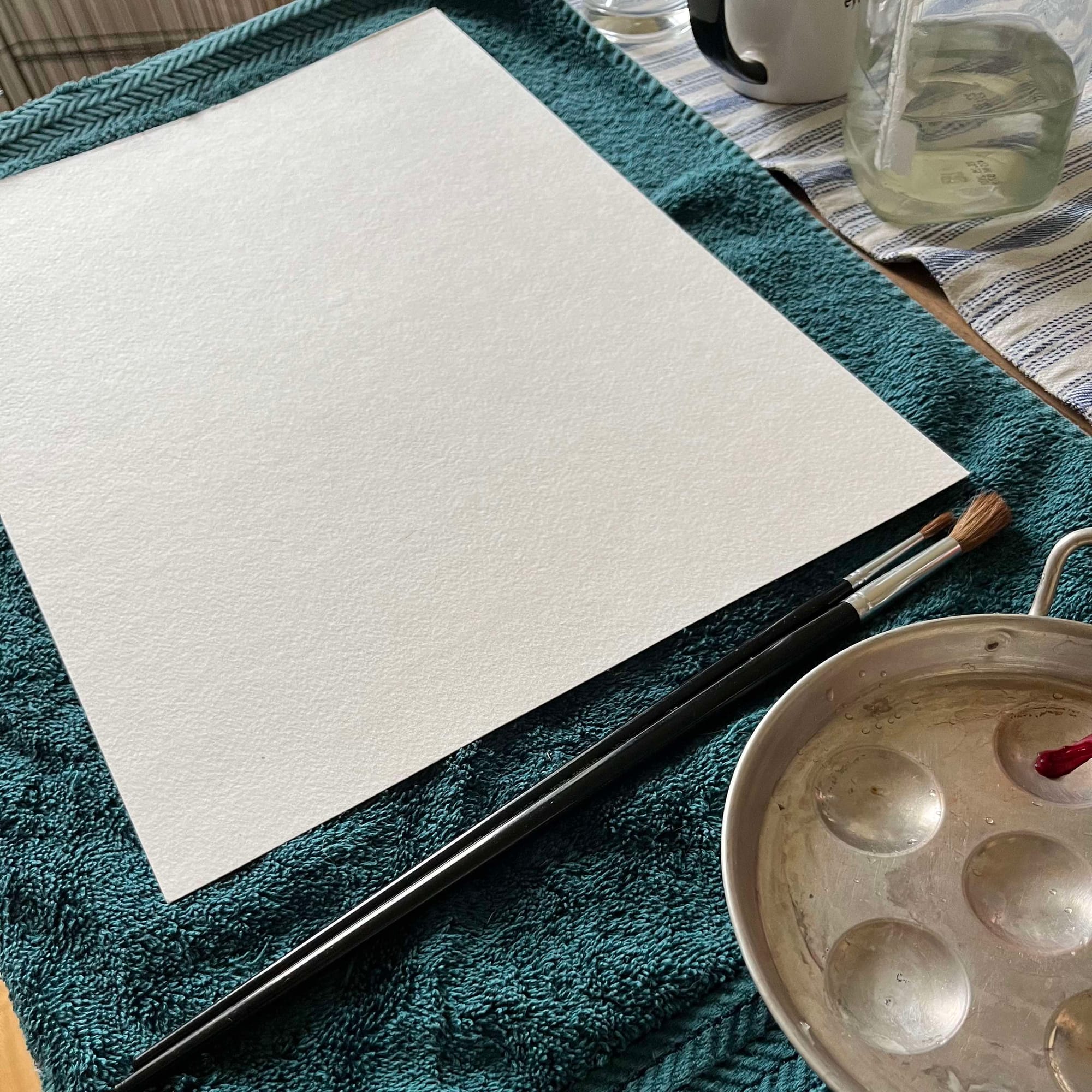 A blank piece of paper, paintbrushes, and a round metal palette