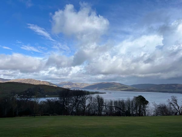 Clouds and blue sky over hills, a bay, a field and trees.