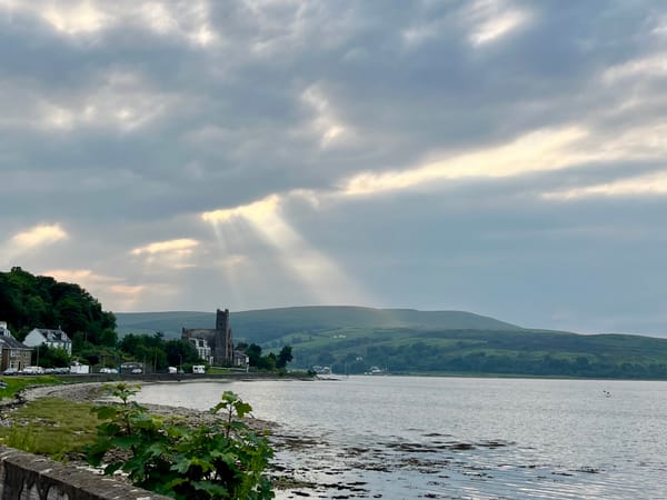 light rays break through clouds above a bay, green hills, and a row of houses